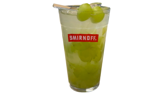 Copo alto com logo vermelho da Smirnoff contendo um coquetel refrescante em tom verde-claro. A bebida está cheia de gelo e uvas verdes cortadas. A borda é decorada com um palito de madeira espetando duas uvas verdes inteiras. Fundo branco isolado.