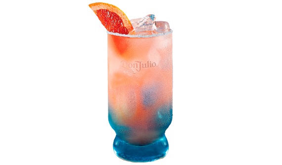 Copo alto suado com base azulada e logo da Don Julio. Contém um coquetel refrescante em tom rosa-coral com gelo. O copo possui uma borda incrustada de sal e é decorado com uma fatia de toranja (grapefruit). Fundo branco.