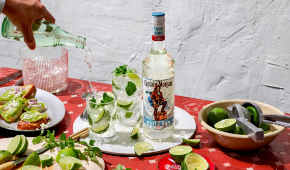 Two highball glasses filled next to Captain Morgan White Bottle with ice, limes and mint