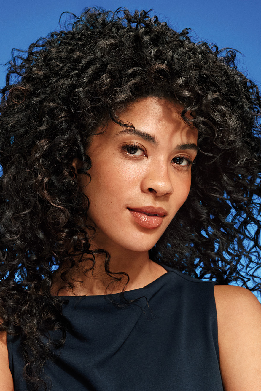 Donna con lunghi capelli neri naturalmente ricci su sfondo blu, che mette in risalto ricci definiti e capelli dall'aspetto sano.