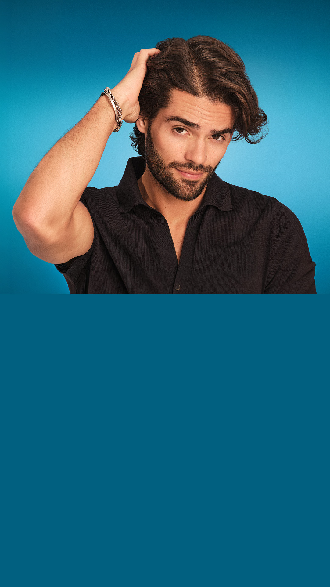 Uomo con capelli scuri e barba che si tocca delicatamente il cuoio capelluto guardando l’obiettivo. Indossa una camicia nera su sfondo blu. L’immagine richiama il comfort del cuoio capelluto e la cura antiforfora.