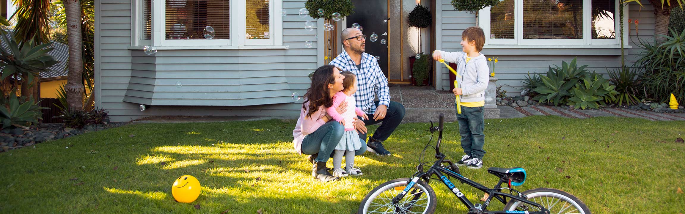 A family playing outside their house in the front yard with a bike, ball and blowing bubbles