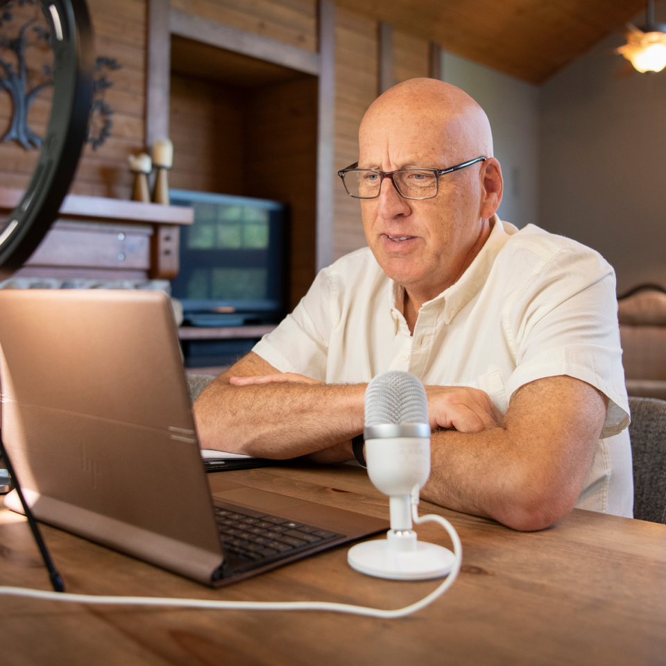 Todd sitting at a table with a laptop, microphone and ring light for a webinar