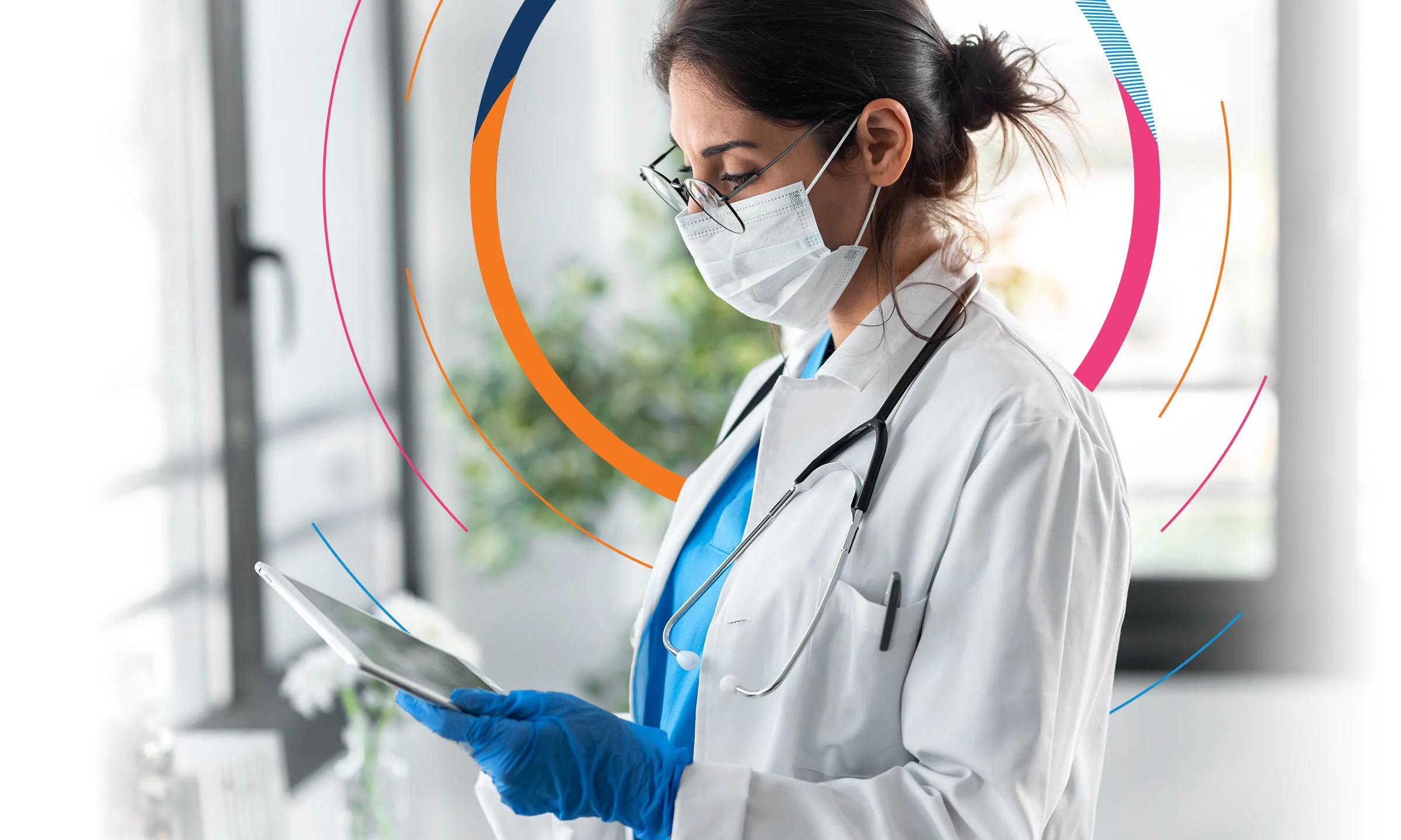 A masked healthcare worker looks down at a tablet 