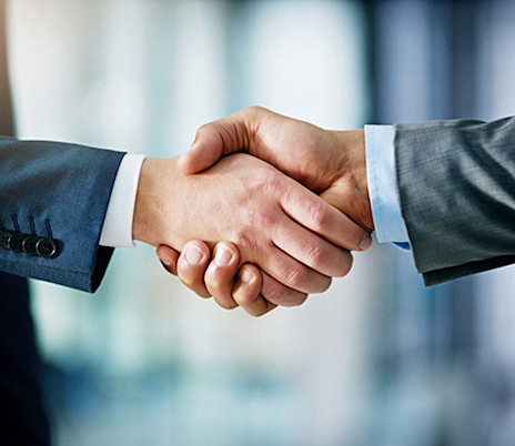 Close-up image of two people shaking hands, both wearing business suits in a professional or office environment. The handshake symbolizes agreement, partnership, and cooperation.