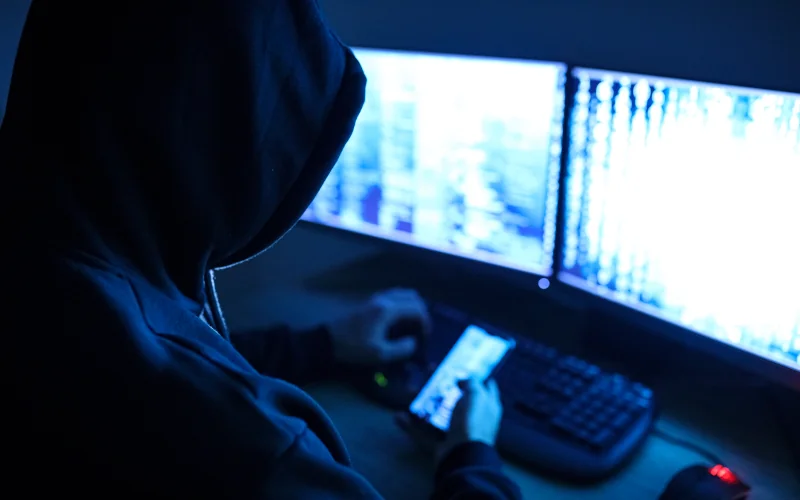 Internetbetrüger sitzt maskiert vor seinem PC und hält ein Handy in der Hand 