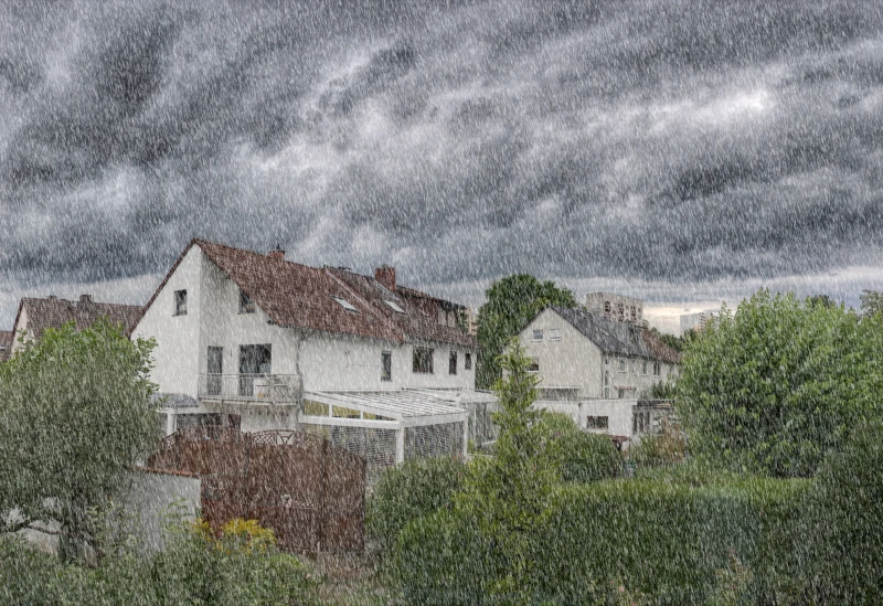 Regen durchströmt die Ansicht auf zwei Hausblockreihen. Die vordere Reihe weist dabei eine rote Bedachung auf während die hintere eine gräuliche kennzeichnet.
