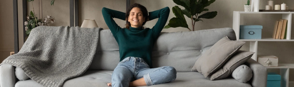 Eine entspannte junge Frau hispanischer Herkunft sitzt in einer entspannten Haltung auf einem großen, bequemen Sofa im Wohnzimmer, atmet tief durch, träumt und lässt ihren Gedanken freien Lauf. Eine erfolgreiche Frau, die gerade eine neue Wohnung gekauft hat, ruht sich zu Hause aus und genießt den Moment.