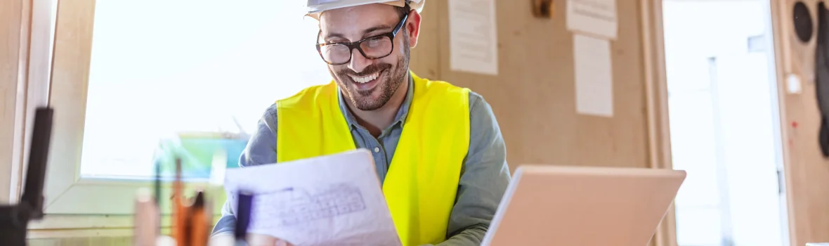 Handwerker sitzt lachend mit Dokumenten in der Hand an einem Laptop