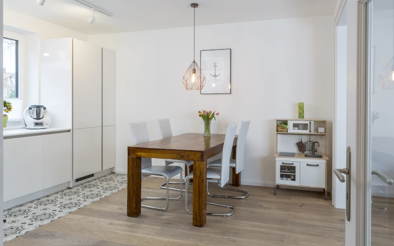Das Bild zeigt einen hellen Essbereich mit Holztisch, weißen Stühlen, moderner Pendelleuchte, angrenzender Küche und einem kleinen Sideboard.