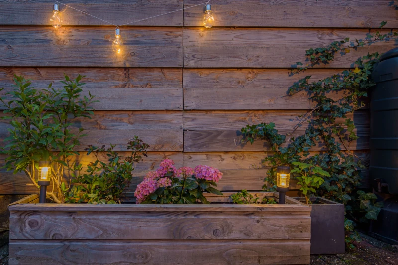Über eine Lichterkette und Lampen wird ein Hochbeet vor einer Holzfassade beleuchtet
