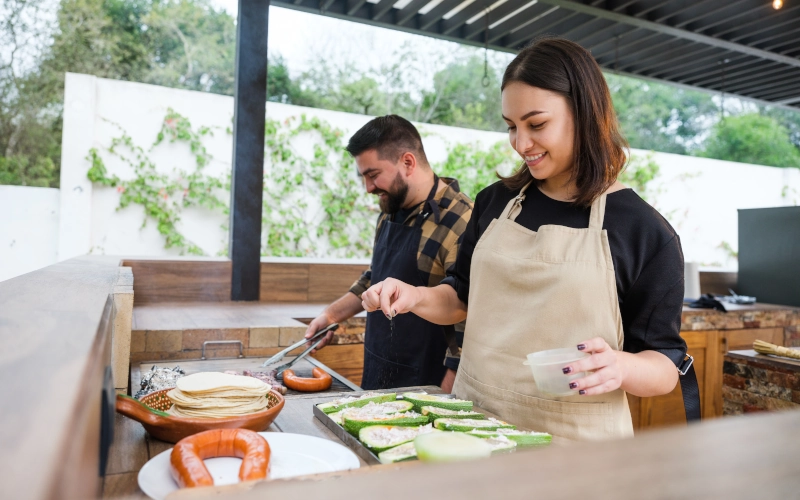 Zwei glücklich aussehende Personen, die zusammen in einer Outdoor-Küche grillen und kochen.