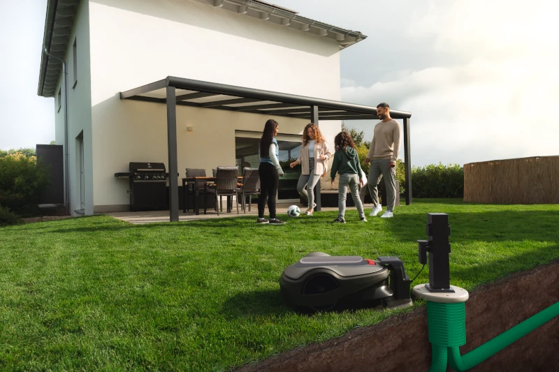 Mähroboter lädt sich im Garten auf, Familie steht auf dem Rasen vor einer Terrasse