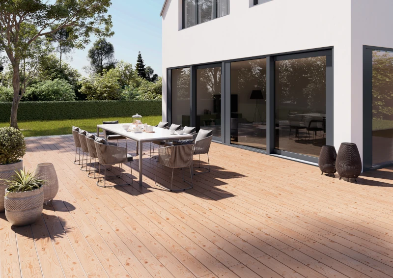 Eine große, sonnenbeschiene Holz-Terrasse vor einem modernen Holzbau mit einer großen Fensterfront. Auf der Terrasse steht ein großer Gartentisch und zehn Stühle.