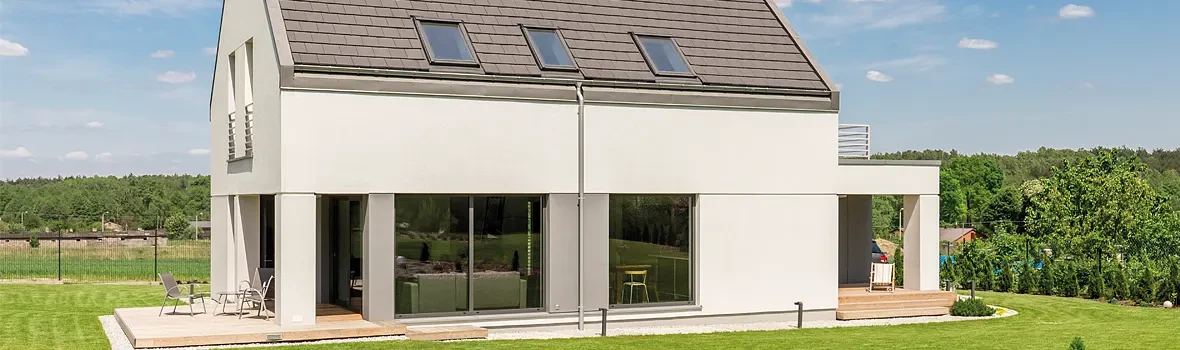 Zu sehen ist ein Haus mit einer großen Fensterfläche, stehend auf einem zwar grünen aber auch kargen Grundstück. Das Haus weißt außerdem eine kleine Holzterrasse auf.