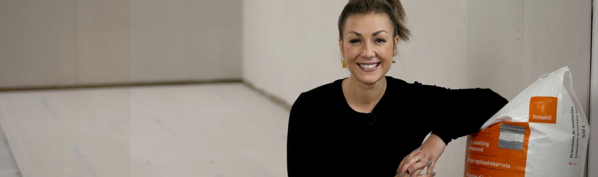 Eine Frau, die in einem Raum auf einen, auf dem Boden liegenden Holzbalken sitzt und sich auch ein Sack mit Ausgleichsschüttung stützt.