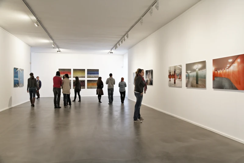 Verschiedene Personen, die in einer Bildgalerie, sich die an der Wand hängenden Bilder, anschauen. Der Boden ist in einer Betonoptik.