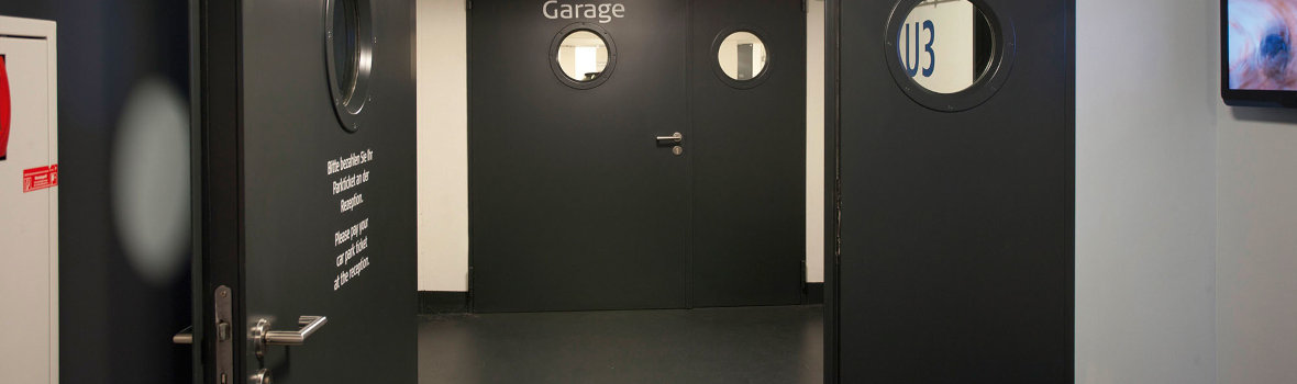 Ein Flur mit schwarzen Teckentrupp Brandschutztüren die zur Garage führen. Türen haben Bullaugen und eine Feuerlöscher Box befindet sich an der Wand.