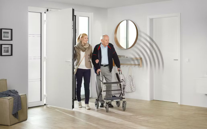 Frau mit älterem Herren am Rollator kommt durch weiße Haustür