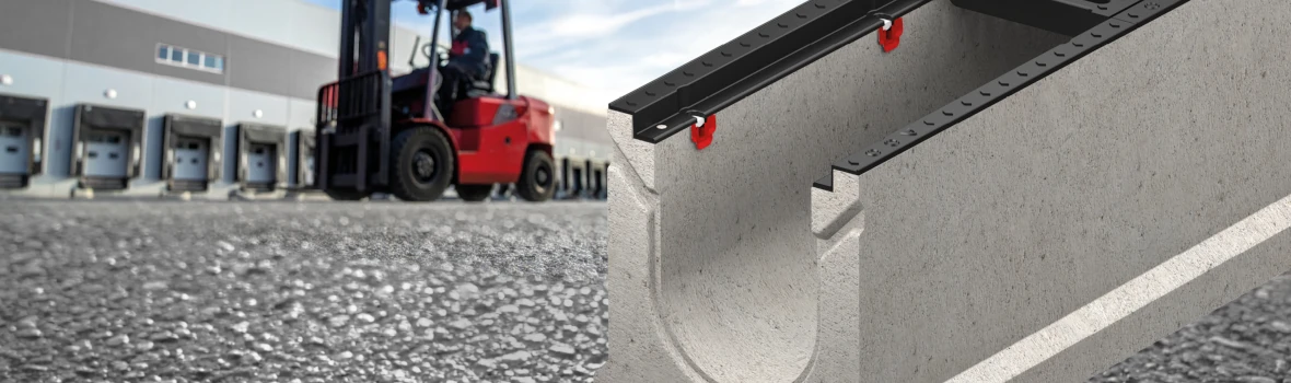 Das Bild zeigt eine Beton-Entwässerungsrinne mit schwarzer Abdeckung auf einem Industriegelände. Im unscharfen Hintergrund sind ein Gabelstapler und Lagerhallen zu sehen, wodurch ein robuster, gewerblicher Einsatzbereich vermittelt wird.