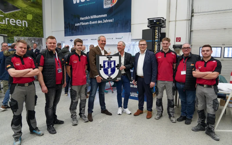 Gruppe von Menschen in einer Halle und zwei Männer in der Mitte halten ein Wappen