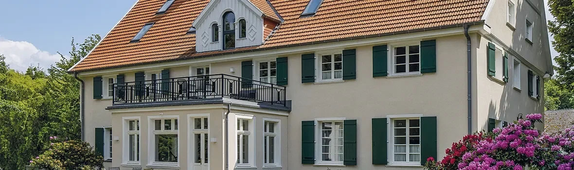 großes beigefarbenes Gebäude mit kleinem Balkon, Fensterläden und Satteldach