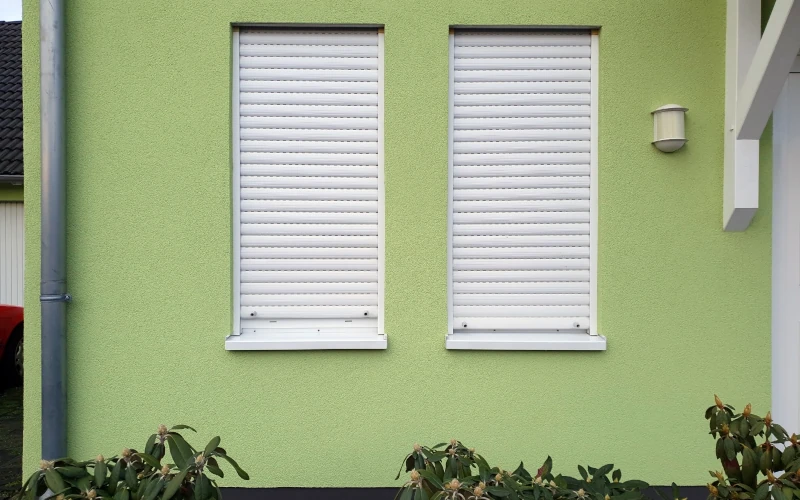Haus mit grüner Putzfassade und zwei durch Außenjalousien geschlossene Fenster