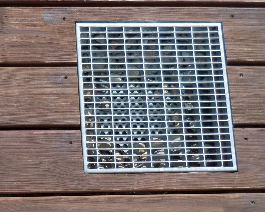 Ein quadratischer Metallgitterrost, eingebettet in eine Fläche aus dunklen Holzplanken. Der Rost dient als Abdeckung für einen darunterliegenden Bereich
