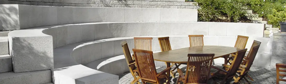 Halbrunde Sitzstufen aus hellem Stein mit Holzsitzgruppe im Mittelpunkt einer Terrasse.