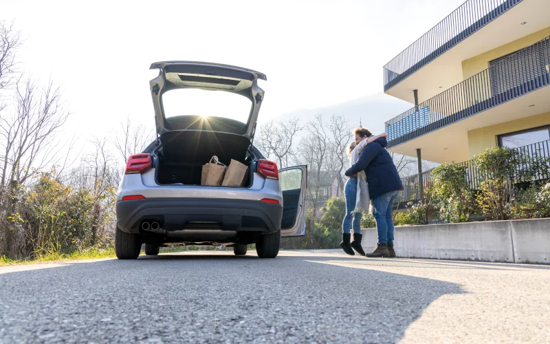 Paar umarmt sich auf einem Hof mit offenem Auto danebenstehend