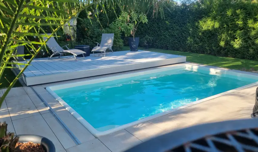 Ein Pool steht in einem Garten. Um ihn herum ist eine Terasse mit hellen Steinen und zwei Liegestühlen.