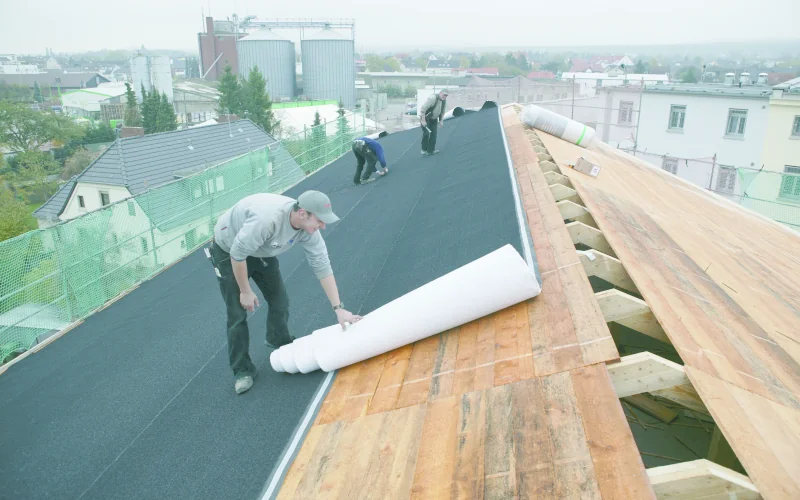 3 Arbeiter legen eine Unterlage auf einem großen Dach aus