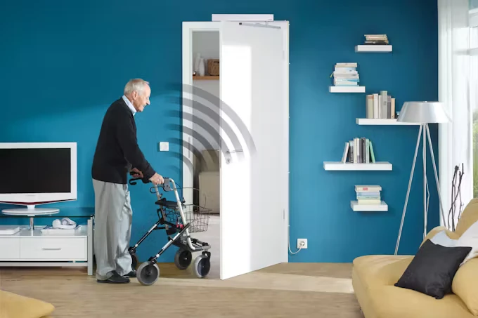 Ein älterer Mann schiebt mit einem Rollator durch eine Wohnungstür, die sich automatisiert öffnet. 