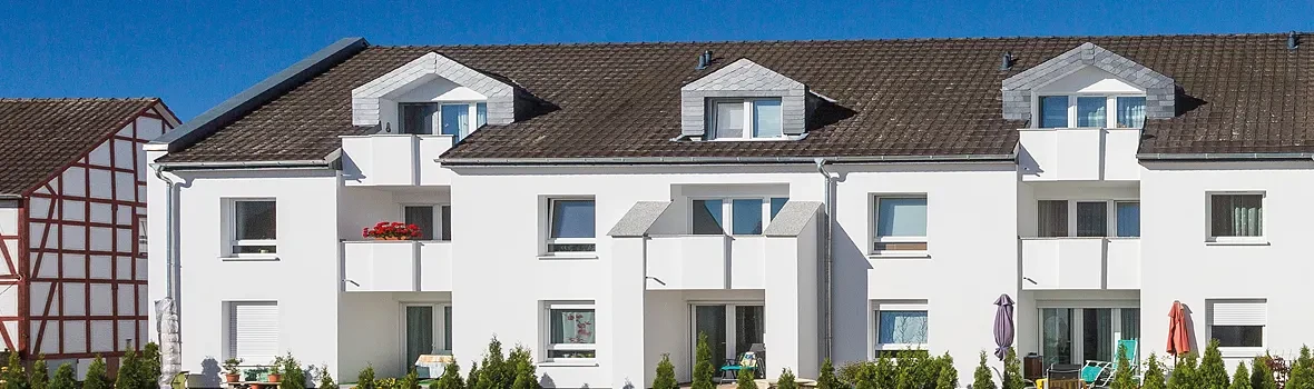 Ein großer, weißer Wohnungskomplex, unterteilt in drei Etagen. Unten befinden sich einzelne Terrassenparzellen, auf den Etagen darüber kleine Balkone.