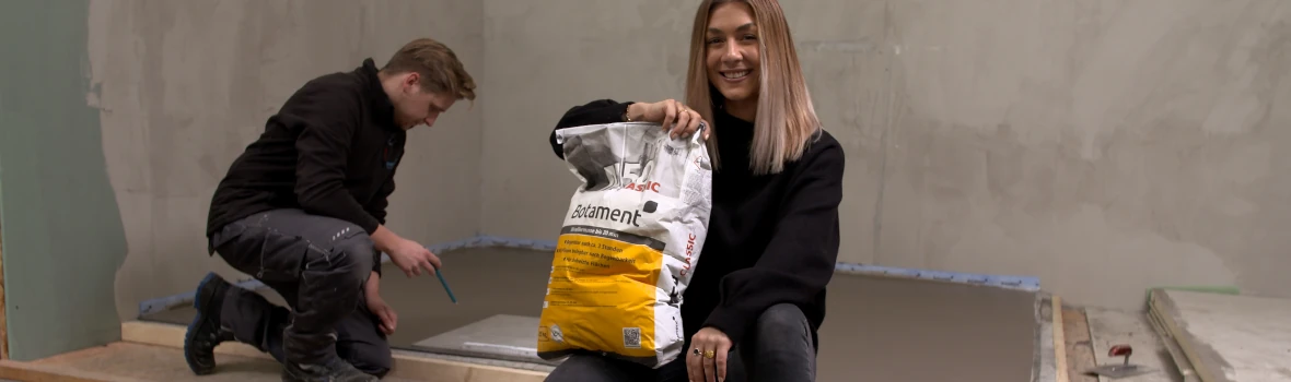 Eine Frau, die hingehockt ein Sack mit Nivelliermasse in der rechten Hand hält und ein Mann im Hintergrund, der auf den Bodenuntergrund schaut.