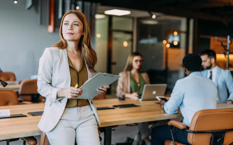 Eine Frau im Business-Outfit steht mit einem Klemmbrett in der Hand in einem modernen Büro und blickt nachdenklich zur Seite, während im Hintergrund mehrere Personen an einem Tisch arbeiten.