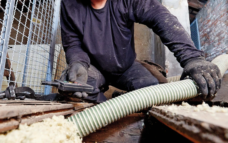 Ein Handwerker bläst mit einem Schlauch Dämmmaterial in eine Bodenöffnung und überprüft dabei die Füllmenge mit einem Messgerät.