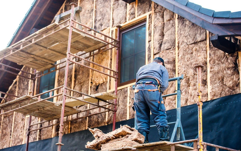 Arbeiter installiert Dämmmaterial an Hausfassade, Gerüst vorhanden, konzentrierte handwerkliche Tätigkeit, Renovierungsprojekt.