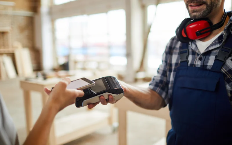 Ein Mann in Handwerkerklamotten hält ein Kartenzahlungsgerät hin. Ein weitere Person bezahlt mit einem Handy.