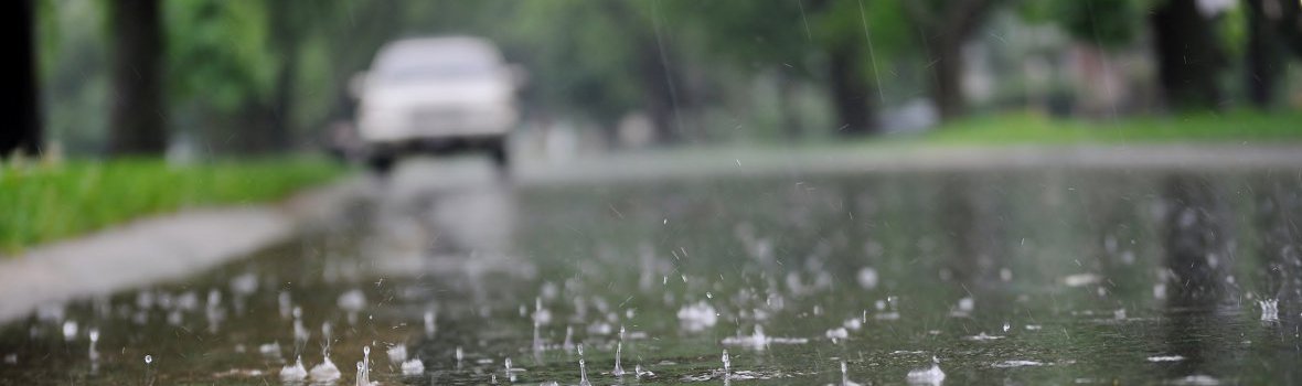 Regentropfen fallen auf Straße, im Hintergrund parkt ein Auto