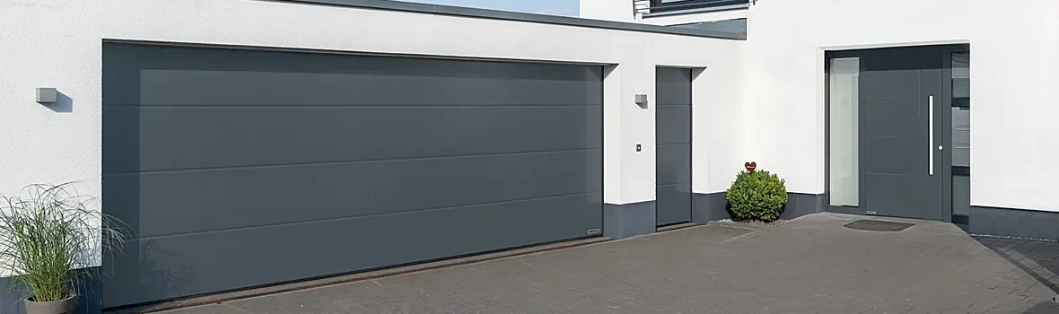 Auf dem Bild zu sehen ist ein anthrazit farbenes Sektionaltor neben einer Haustür. Die Fassade ist weiß.
