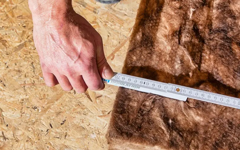 Hand misst Dämmmaterial mit einem Zollstock auf Holzplatte.