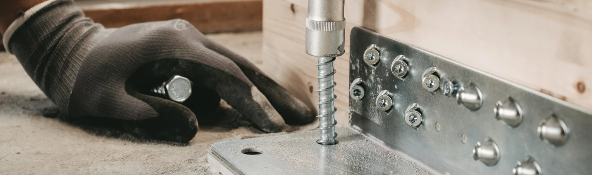 Mit einer großen Schraube wird mit einem aufwendigen Metall-Winkel ein Holzelement mit einer Betonfläche verbunden.