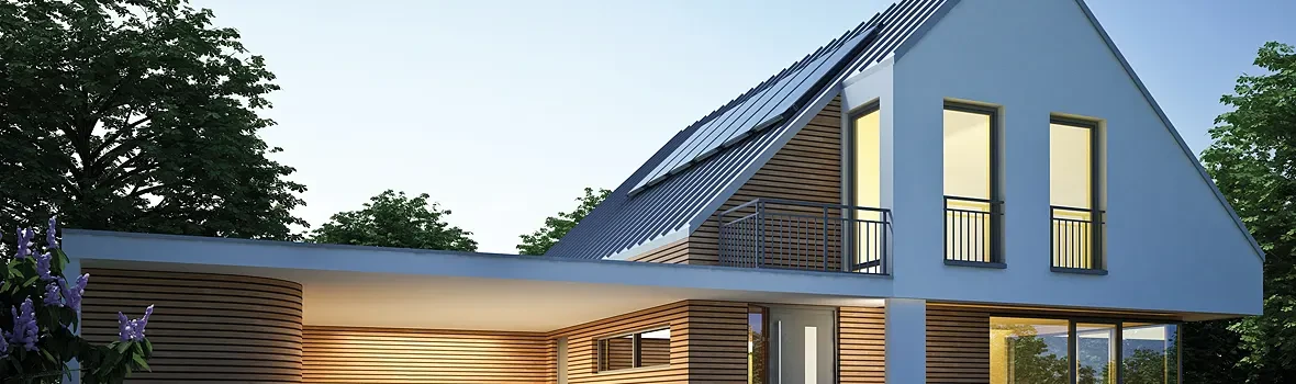 Ein Haus mit Holzelementen an der Fassade ist umgeben von Bäumen in der Dämmerung. Im Haus selber brennt Licht.