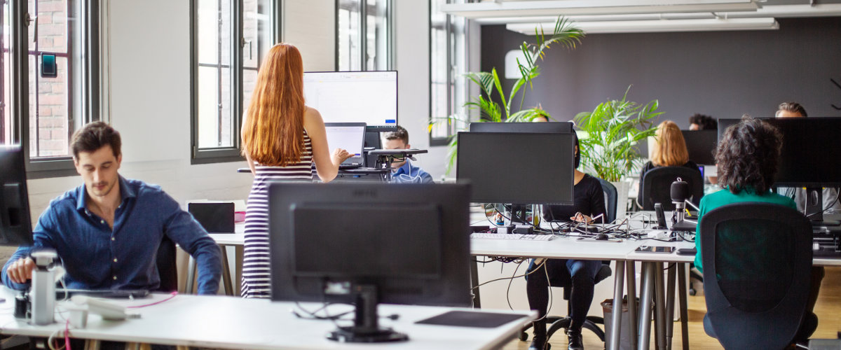 Ein modernes Büro mit mehreren Personen, die an Computern arbeiten. Eine Frau mit langen roten Haaren steht und arbeitet an einem höhenverstellbaren Schreibtisch. Im Hintergrund sind große Fenster und einige Pflanzen zu sehen.