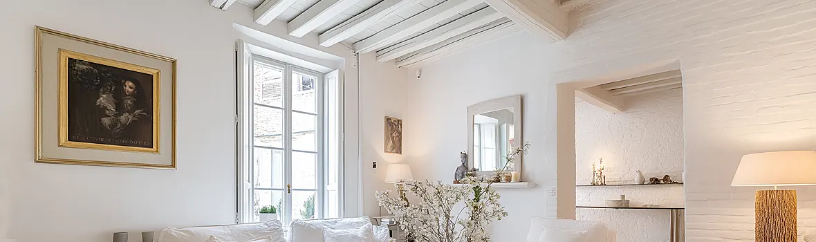 Das Bild zeigt einen hellen, stilvoll eingerichteten Wohnraum in Weiß- und Cremetönen mit großem Fenster, sichtbaren Deckenbalken und eleganten Dekoelementen. Insgesamt wirkt der Raum ruhig, hochwertig und leicht mediterran.