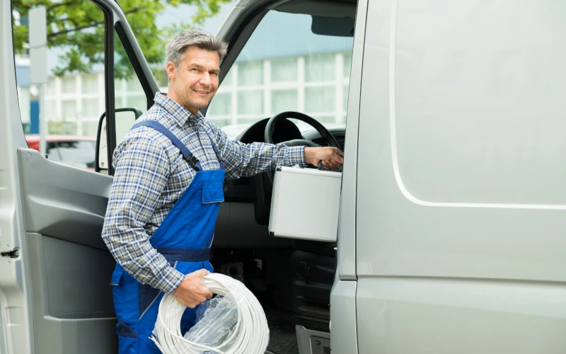 Ein Handwerker in blauem Overall steht an einem Lieferwagen und hält ein Kabel, während er lächelt.
