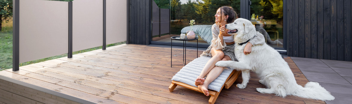 Frau sitzt auf Liege mit Hund auf Holzterrasse mit Sichtschutzelementen