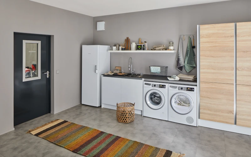 Hauswirtschaftsraum mit Brandschutztür, Kühlschrank und Arbeitsfläche mit Waschbecken,  Waschmaschine und Trockner, daneben ein Schrank. 