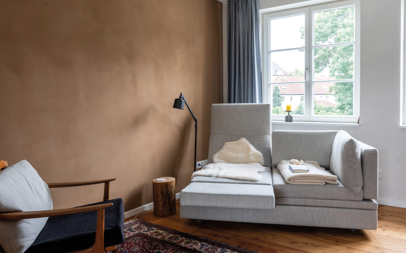 Wohnzimmer mit brauner Wand, grauem Sofa, Sessel, Stehlampe und Fenster mit Vorhang.